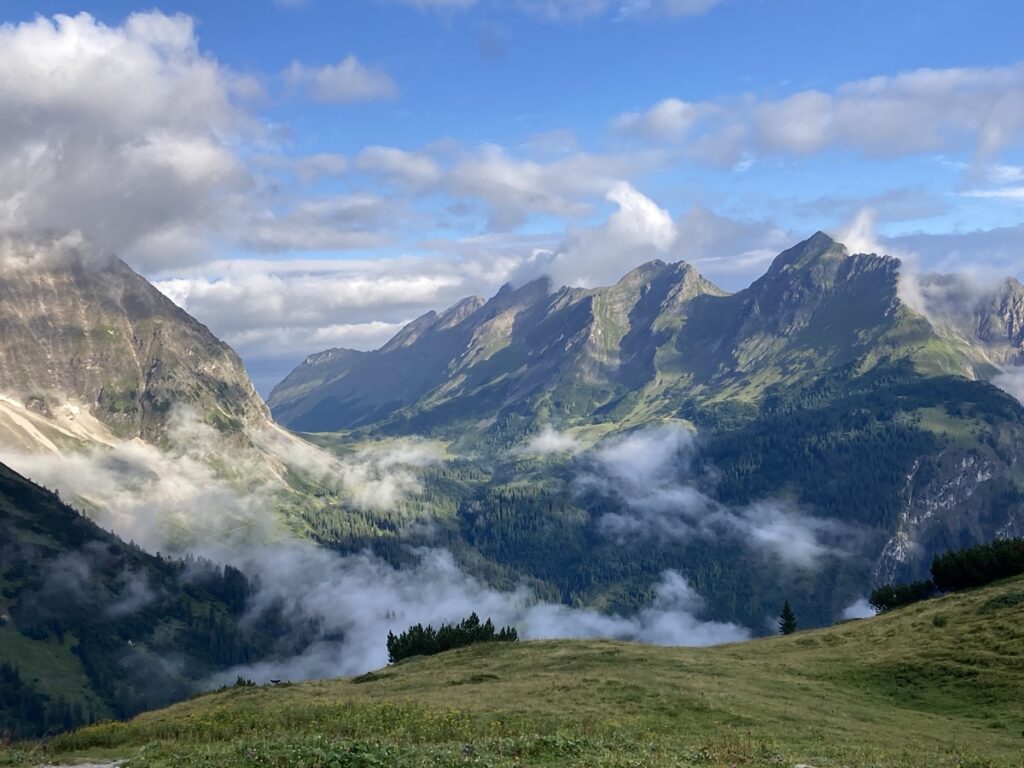 Gebirgskette mit zerklüfteten Gipfeln, vereinzelten Wolken und Sonnenlichtflecken über einem grünen Tal mit Bäumen und grasbewachsenem Vordergrund.