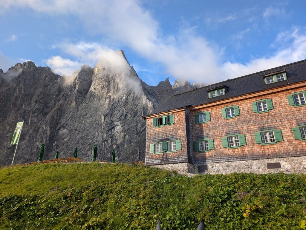 Ein Holzgebäude mit grünen Fensterläden steht auf einem grasbewachsenen Hügel vor felsigen, wolkenverhangenen Bergen unter blauem Himmel.