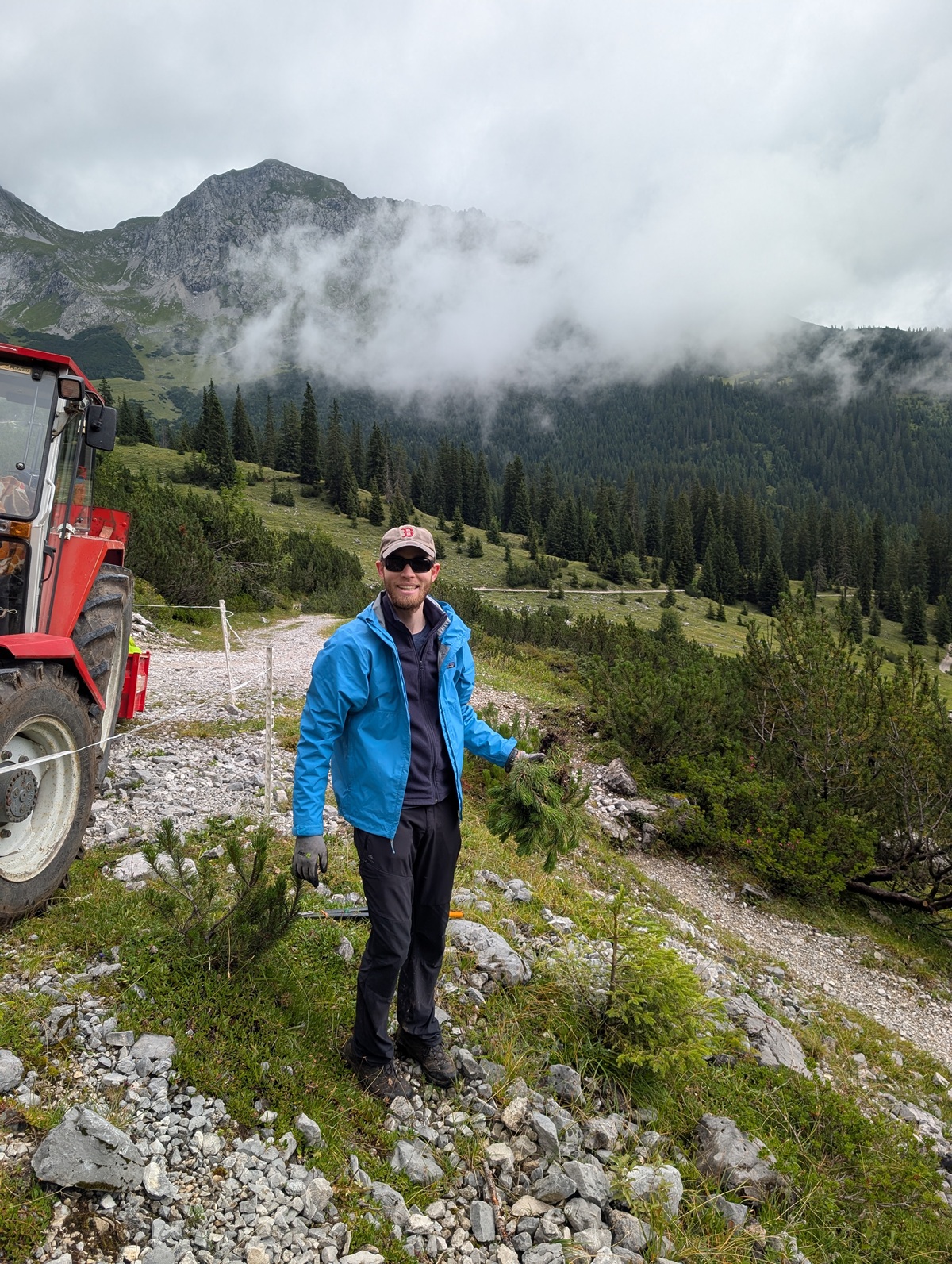 Ein Mann in blauer Jacke und Mütze steht auf einem felsigen Pfad in einer Berglandschaft mit Bäumen, Wolken und einem roten Traktor in der Nähe.
