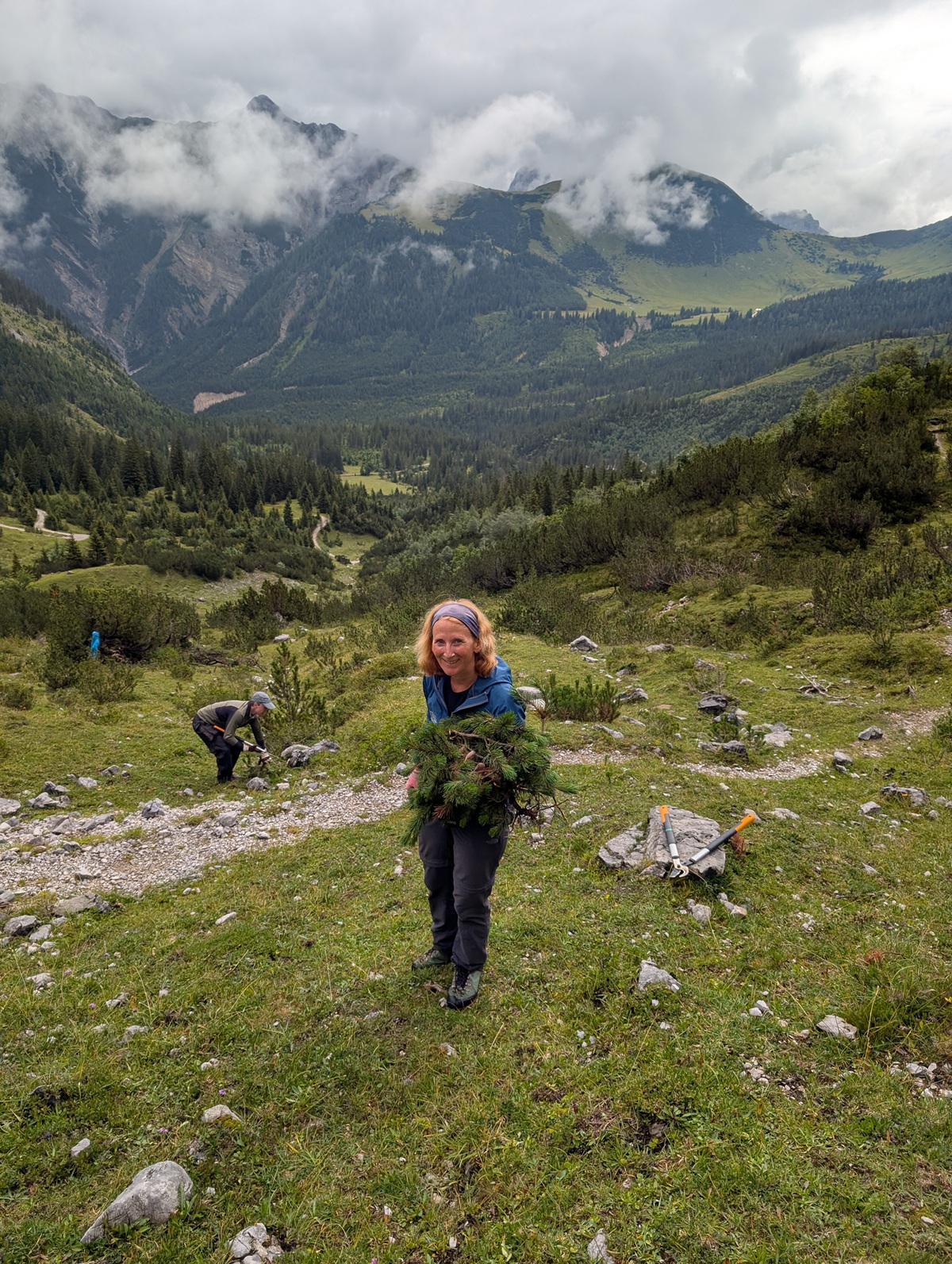 Eine Person hält ein Bündel grüner Zweige, während sie an einem bewölkten Tag auf einem felsigen Pfad in einer bergigen, bewaldeten Landschaft steht; eine andere Person arbeitet im Hintergrund.