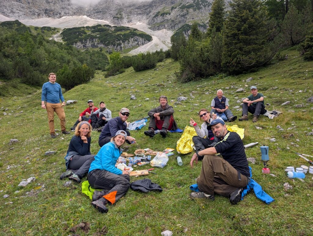 Eine Gruppe von Menschen sitzt im Gras und macht ein Picknick in einer bergigen Gegend mit felsigen Hängen und Bäumen im Hintergrund.