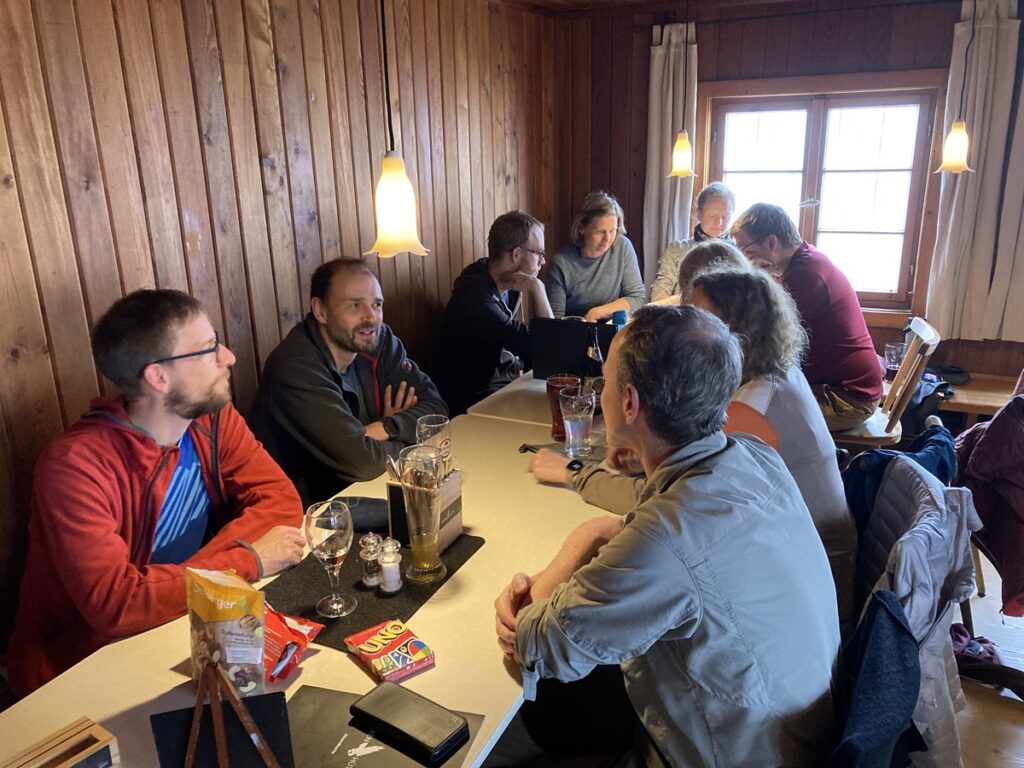 Eine Gruppe von Menschen sitzt in einem hölzernen Raum um einen langen Tisch herum und unterhält sich mit Getränken und Snacks auf dem Tisch.