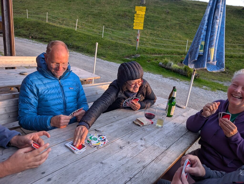 Vier Personen sitzen an einem hölzernen Picknicktisch im Freien auf der Almpflege Martinsbergalm und spielen ein Kartenspiel mit Getränken in der Nähe; eine Person legt eine Karte auf den Stapel, während die anderen zusehen und lachen.