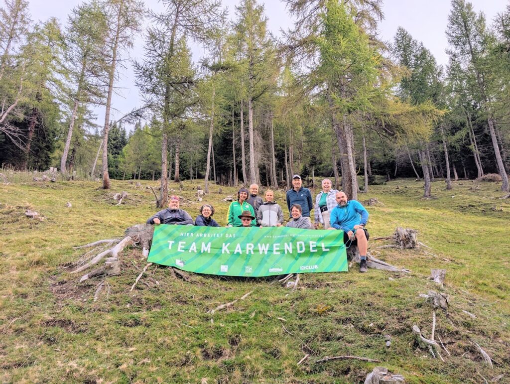 Eine Gruppe von Menschen auf einem grasbewachsenen Hügel bei der Almpflege Martinsbergalm, die ein grünes Banner mit der Aufschrift "Team Karwendel" halten, umgeben von Bäumen.