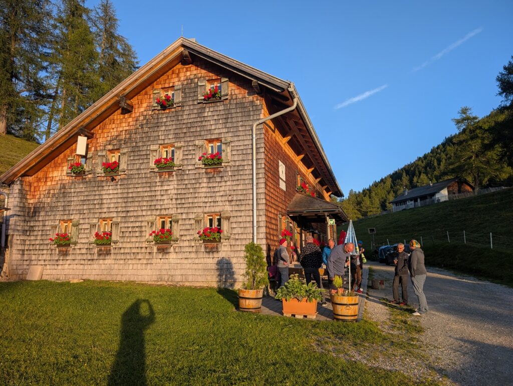 Eine Gruppe von Menschen versammelt sich vor dem rustikalen Holzhaus der Almpflege Martinsbergalm, mit Blumenkästen an den Fenstern, umgeben von Gras und Bäumen im Abendsonnenlicht.