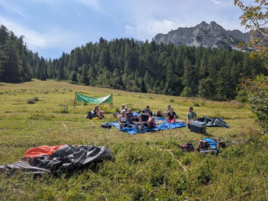 Eine Gruppe von Menschen sitzt auf Planen und macht ein Picknick auf einer grasbewachsenen Lichtung, umgeben von Campingausrüstung und bewaldeten Bergen - der perfekte Ort für ein Treffen der Biotoppflege Issanger.