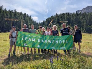 Eine Gruppe von Menschen steht im Freien auf einer Wiese und hält ein grünes "Team Karwendel"-Transparent während der Biotoppflege Issanger, mit Bäumen und Bergen im Hintergrund.