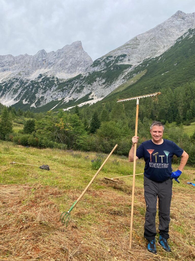 Ein Mann in Outdoor-Kleidung steht mit einem großen Rechen auf einer grasbewachsenen Bergwiese und nimmt an der Biotoppflege Issanger teil, mit Kiefern und felsigen Bergen im Hintergrund.