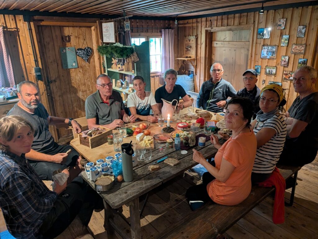 Zehn Personen sitzen um einen Holztisch in einer rustikalen Hütte und teilen sich eine Mahlzeit mit verschiedenen Speisen und Getränken. Die Atmosphäre ist ungezwungen und der Raum ist warm beleuchtet.