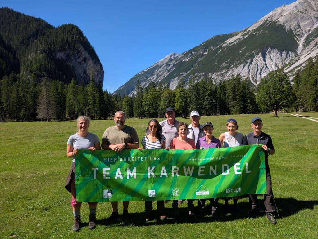 Acht Personen stehen im Freien auf einer Wiese vor Bergen und Bäumen und halten ein grünes Transparent mit der Aufschrift "TEAM KARWENDEL".