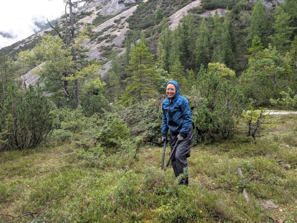 Eine Person in blauer Regenjacke und dunkler Hose steht in einer grünen, bewaldeten Berglandschaft und hält Wanderstöcke in der Hand.