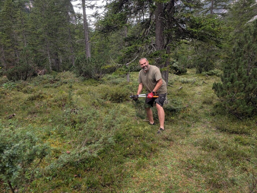 Ein Mann steht auf einer Waldlichtung, hält eine Schaufel in der Hand und lächelt in die Kamera. Er trägt ein T-Shirt, Shorts und Sandalen. Dichte Bäume und Grünflächen umgeben ihn.