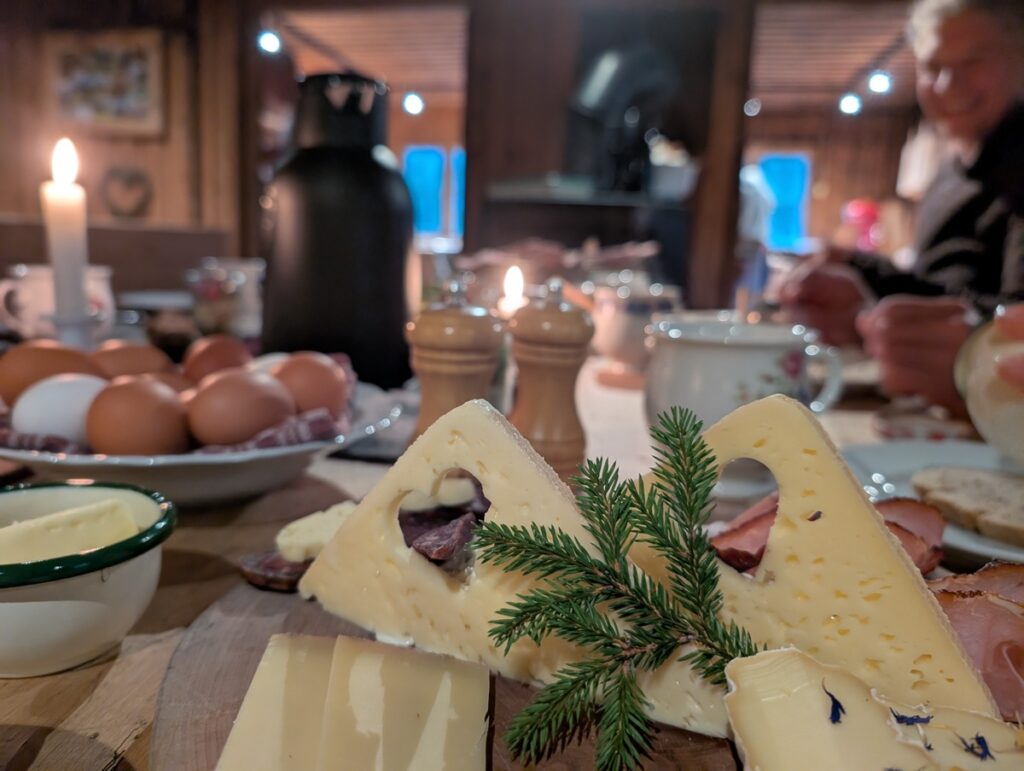 Ein Holztisch, gedeckt mit verschiedenen Käsesorten, Eiern, Butter und Geschirr. Ein kleiner immergrüner Zweig schmückt den Käse. Kerzenlicht und unscharfe Menschen schaffen eine gemütliche Atmosphäre im Haus.