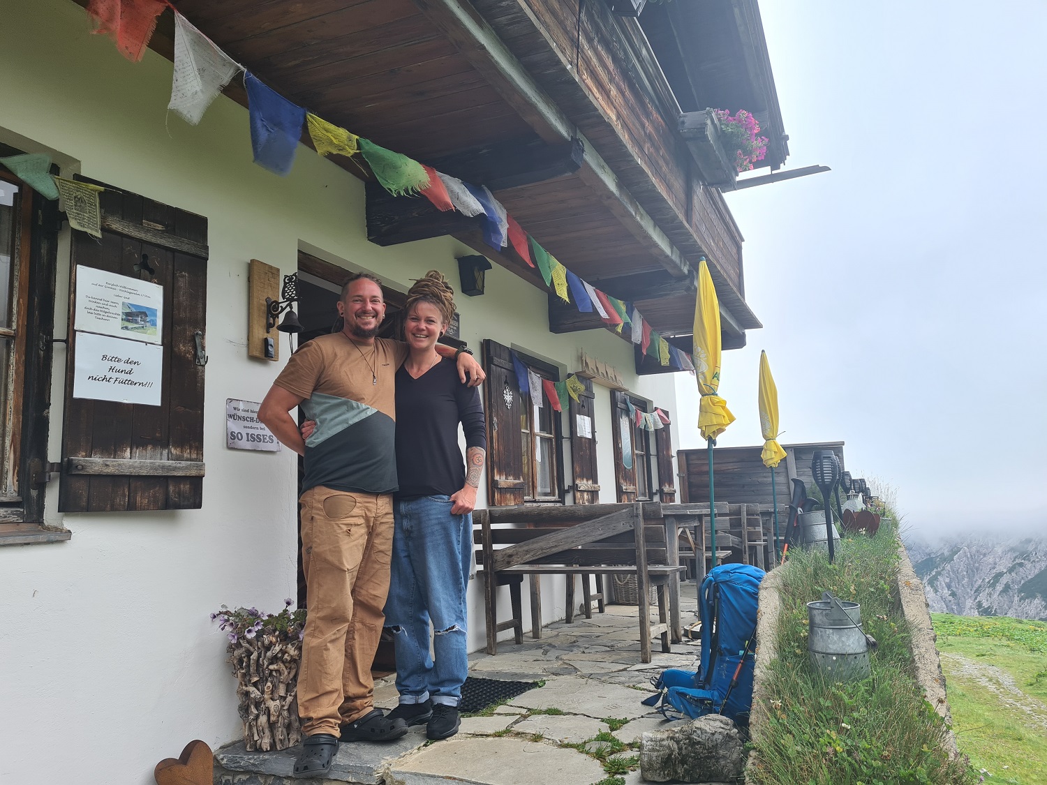 Zwei Menschen stehen lächelnd vor einer rustikalen Berghütte mit bunten Gebetsfahnen, Sitzgelegenheiten im Freien und einer nebligen Landschaft - ein perfekter Ort für KlimaPädagogik-Gespräche inmitten der Schönheit der Natur.