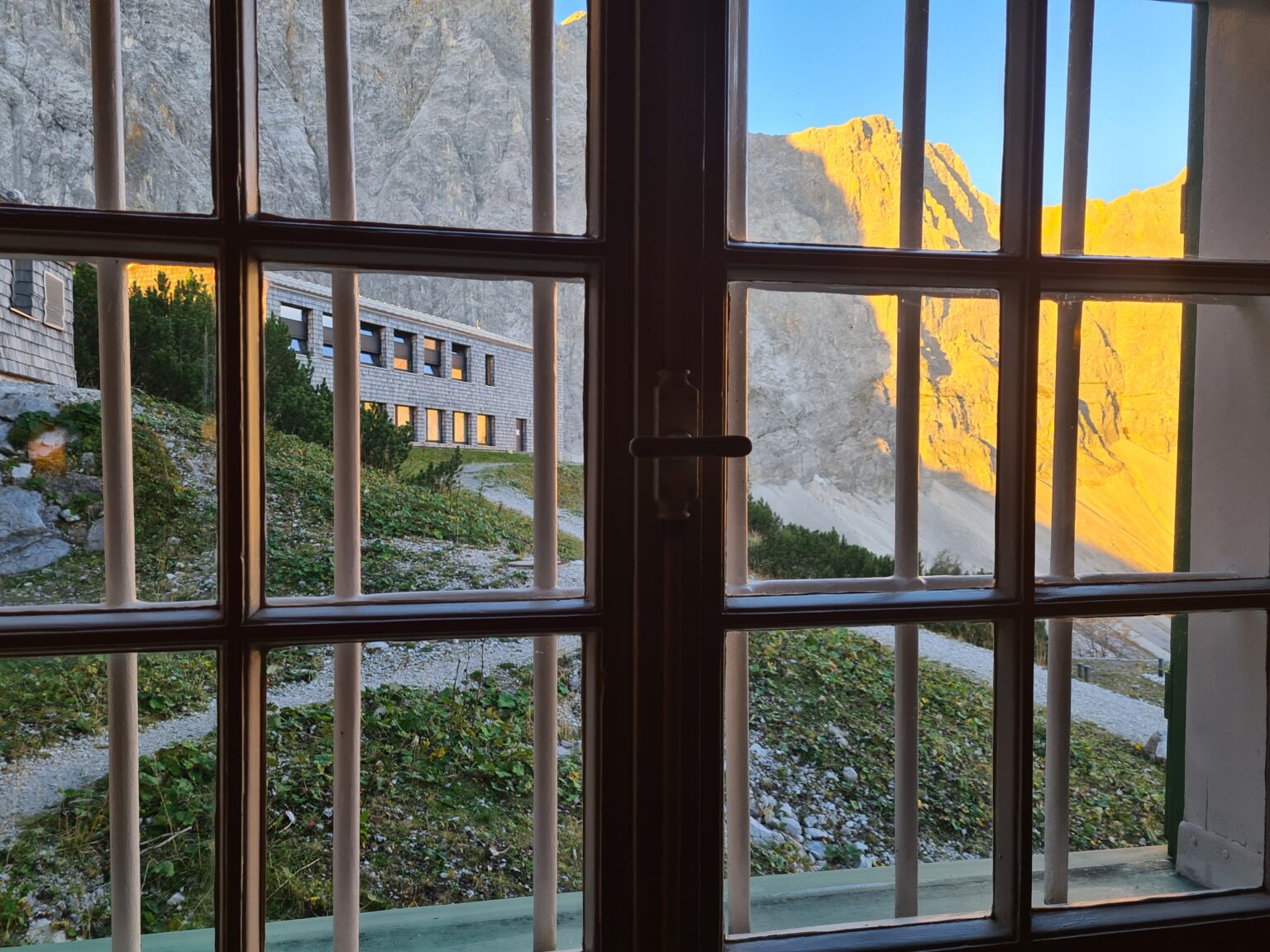 Der Blick durch ein Fenster mit Gitterscheiben offenbart die herrliche Berglandschaft der Kastenalm mit einem sonnenbeschienenen Weg und einem Gebäude inmitten von Sonnenlicht und Schatten.