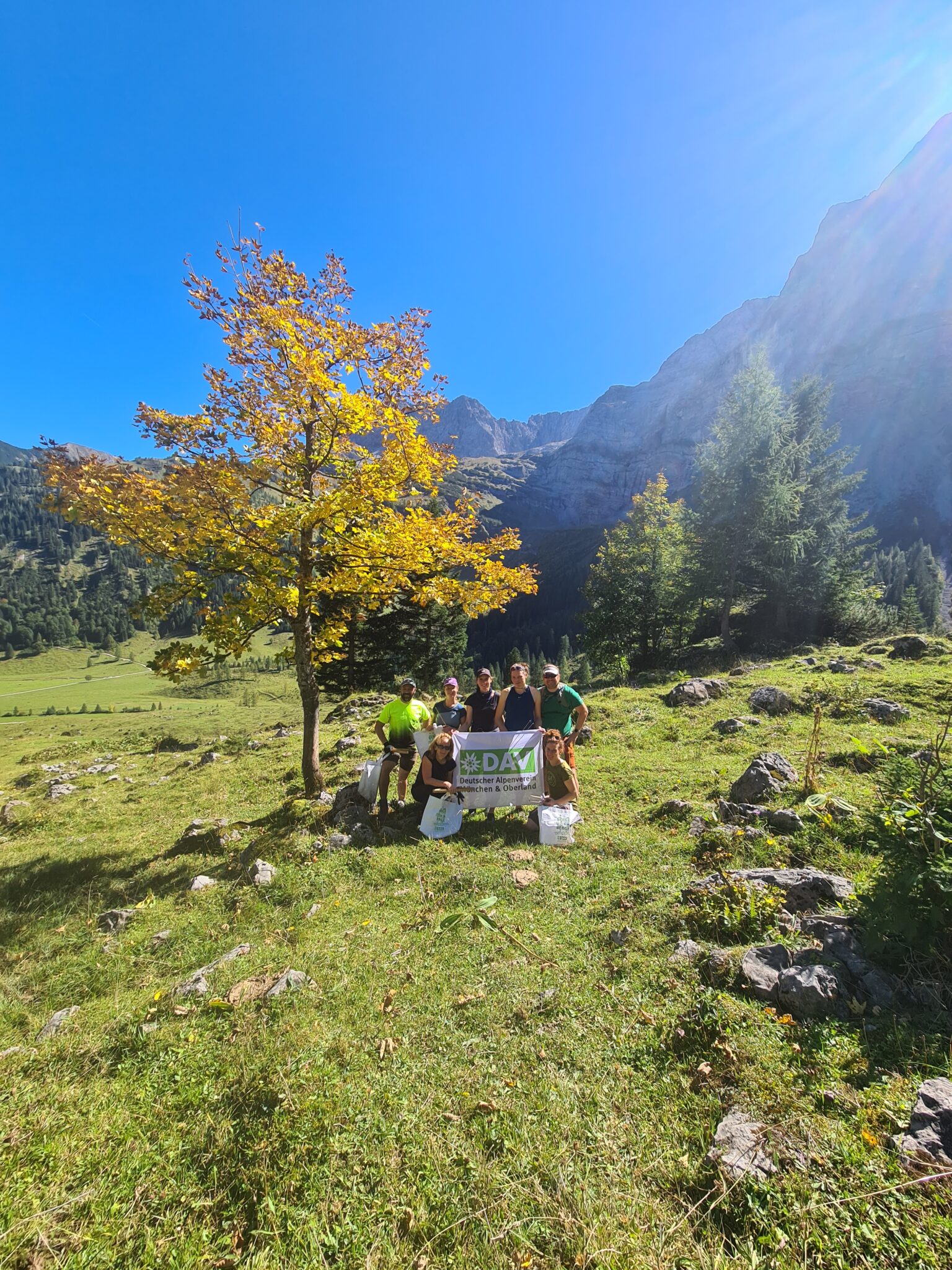 Fünf Personen posieren hinter einem Kastenalm-Banner auf einer Wiese mit Felsen, einem Baum und Bergen im Hintergrund unter einem klaren blauen Himmel.