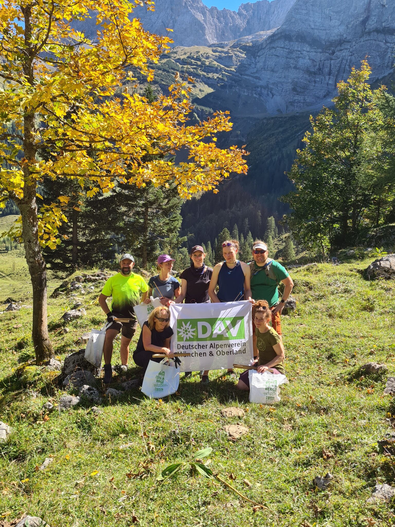 Sieben Personen posieren im Freien auf einem grasbewachsenen Hügel in der Nähe der Kastenalm mit Bergen im Hintergrund und halten ein Banner des Deutschen Alpenvereins München & Oberland".