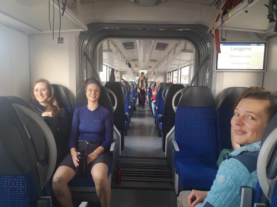 Drei Personen sitzen und lächeln in einem Zugwaggon mit blauen Sitzen. Auf einem Schild über der Tür steht unter anderem "Lenggries" und Kastenalm. Im Hintergrund sind andere Fahrgäste zu sehen.