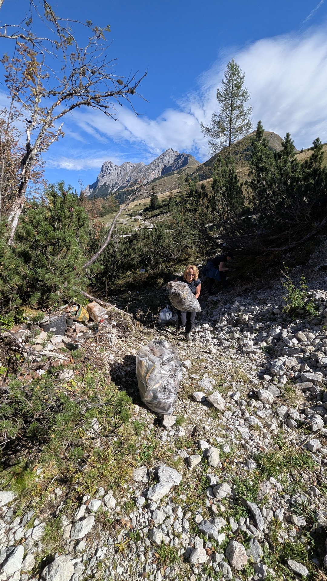 Eine Person sammelt in der felsigen, bergigen Landschaft der Kastenalm bei klarem Himmel Müll ein. Im Vordergrund sind ferne Gipfel und gefüllte Müllsäcke zu sehen.
