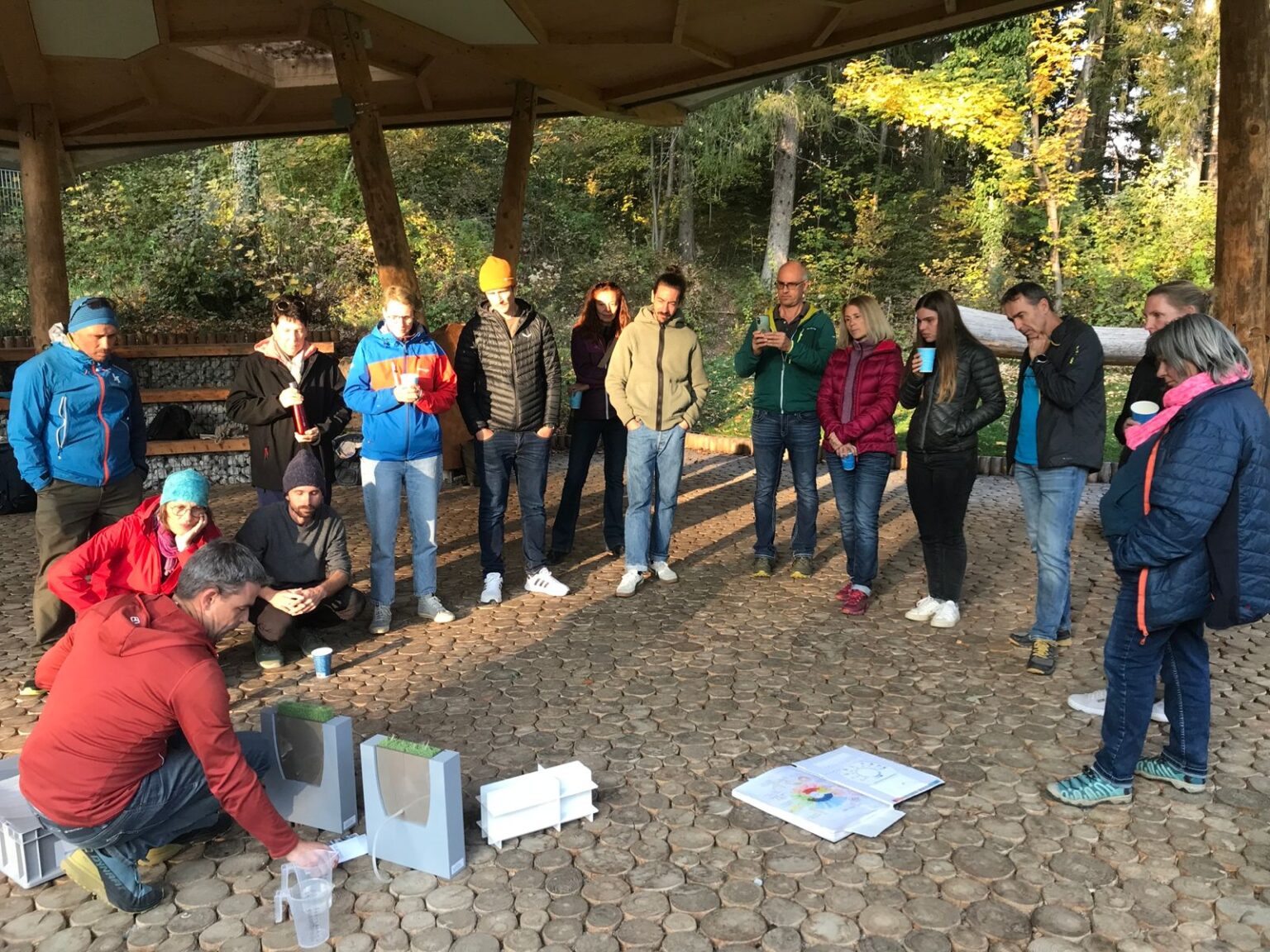 Eine Gruppe von Menschen steht in einem Halbkreis unter einem Unterstand und beobachtet eine Person, die auf dem Boden kniet und die Klimaausrüstung vorführt.