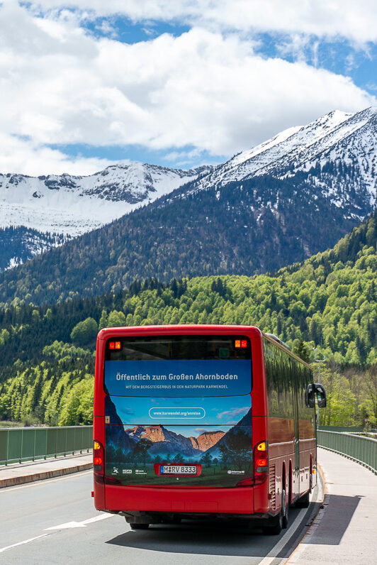 Ein roter Bus fährt die landschaftlich reizvolle Anreise über eine Straße an einem See entlang, umrahmt von grünen Wäldern und schneebedeckten Bergen unter einem teilweise bewölkten Himmel.