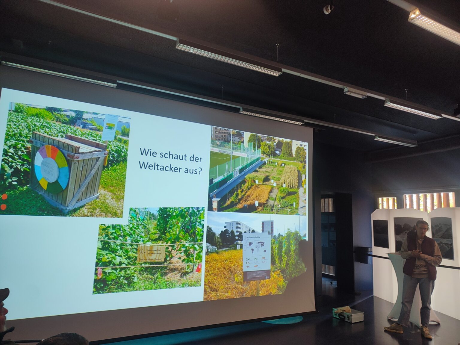 Eine Präsentationsfolie zeigt vier Fotos eines Schrebergartens und eine kreisförmige Grafik, die Klimaaspekte hervorhebt, mit dem deutschen Text "Wie schaut der Weltacker aus?". Zwei Personen stehen neben der projizierten Leinwand.