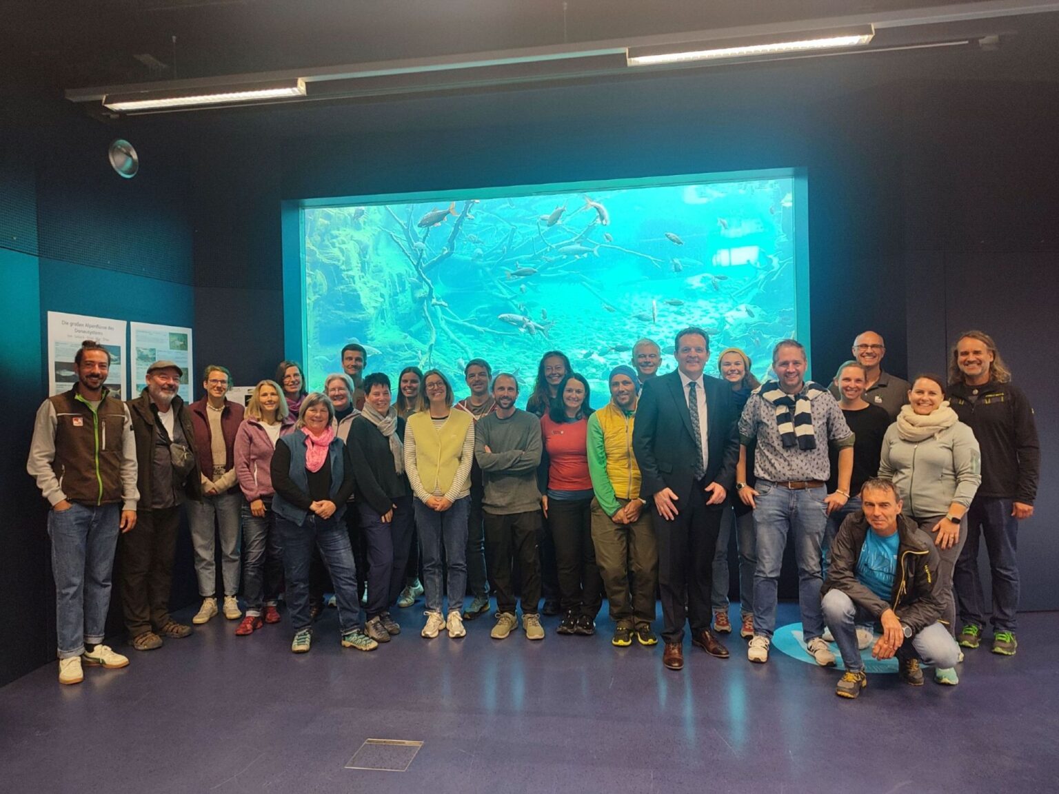 Eine Gruppe von 25 Personen posiert für ein Foto vor einem großen Aquarium und genießt das kühle Raumklima.