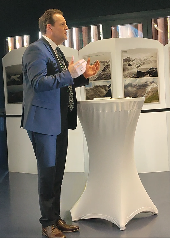 Umwelt-Landesrat René Zumtobel in einem blauen Anzug steht und gestikuliert, während er an einem runden, mit einem weißen Tuch bedeckten Tisch spricht, an dem im Hintergrund Landschaftsfotos von Klima zu sehen sind.