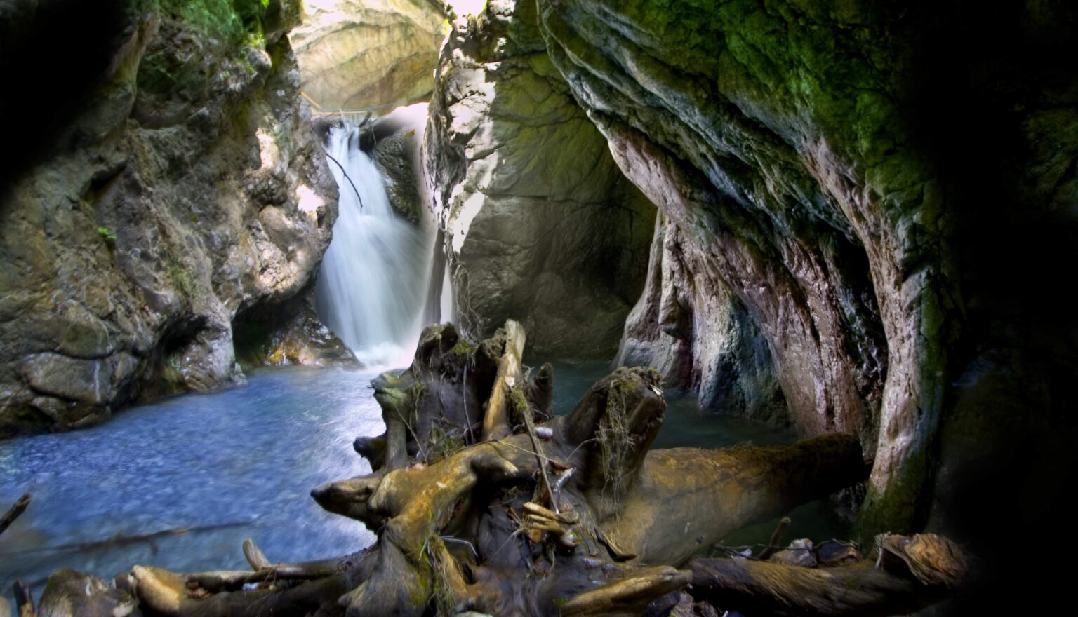 Ein kleiner Wasserfall fließt in ein blaues Becken, das von schroffen Felsen und großen Treibhölzern umgeben ist und an die landschaftliche Schönheit erinnert, die man auf einer Reither Spitze Bergtour durch bewaldete Höhlen und Schluchten findet.