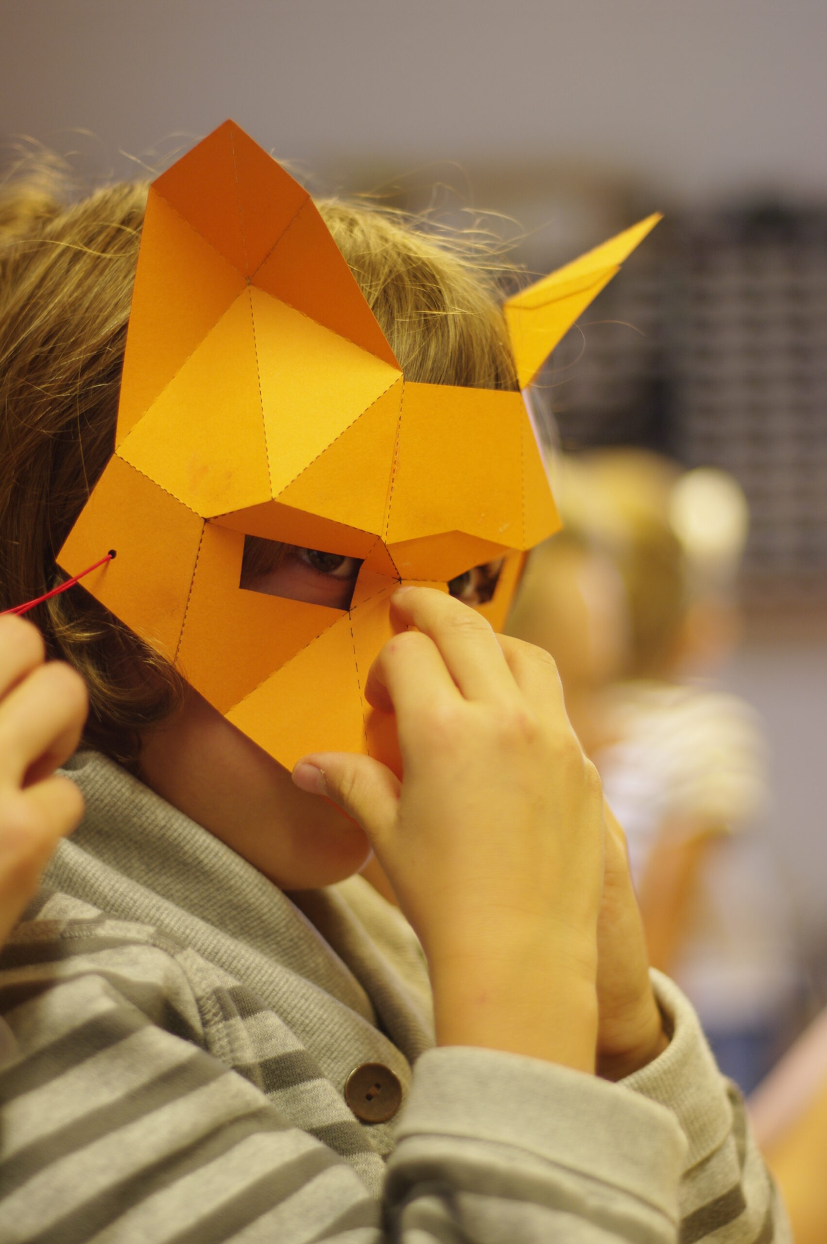 Ein Kind in einem gestreiften Hemd setzt sich eine orangefarbene, geometrische Tiermaske auf, bereit, sich auf ein Junior-Ranger-Abenteuer einzulassen.