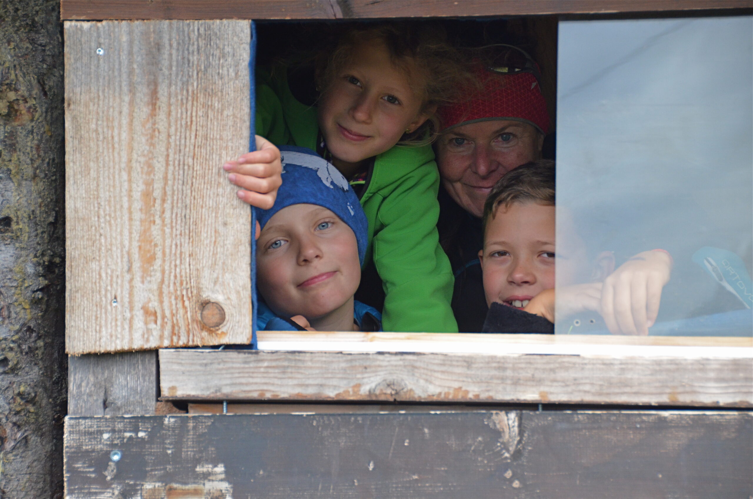 Vier Personen, darunter drei Kinder und ein Erwachsener, schauen aus einem hölzernen Fenster, lächeln wie Junior-Ranger und lehnen sich eng aneinander in einem rustikalen Gebäude.