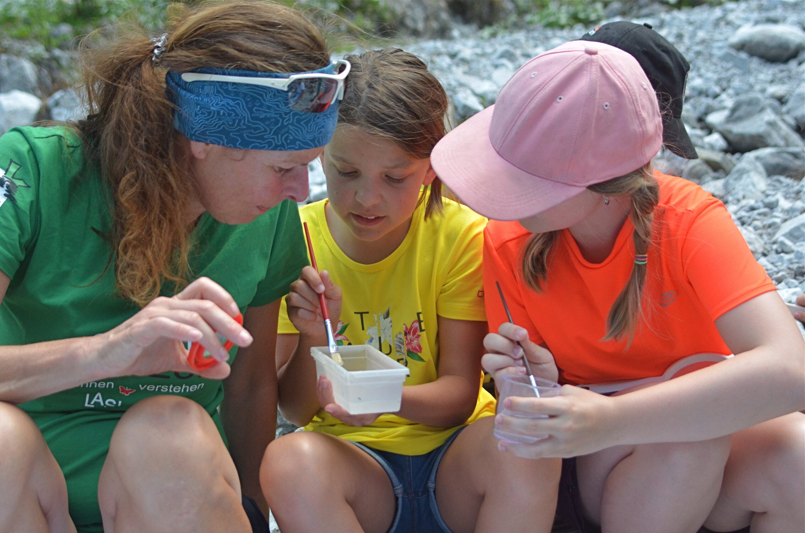 Drei Personen sitzen im Freien auf Felsen und untersuchen Objekte in Plastikbehältern mit Stöcken und Werkzeugen, um an einer Junior-Ranger-Aktivität teilzunehmen.