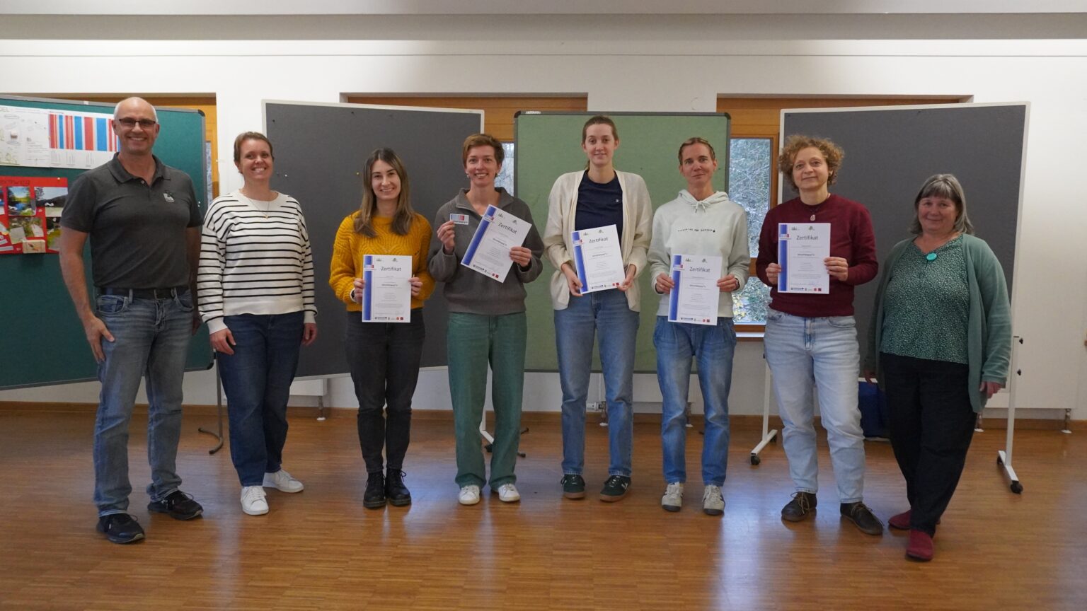 Acht Personen stehen in einem Innenraum; fünf in der Mitte halten Urkunden und lächeln in die Kamera, während drei KlimaPädagog*innen und andere zu beiden Seiten stehen und ebenfalls lächeln. Hinter ihnen ist ein Klassenzimmer zu sehen.