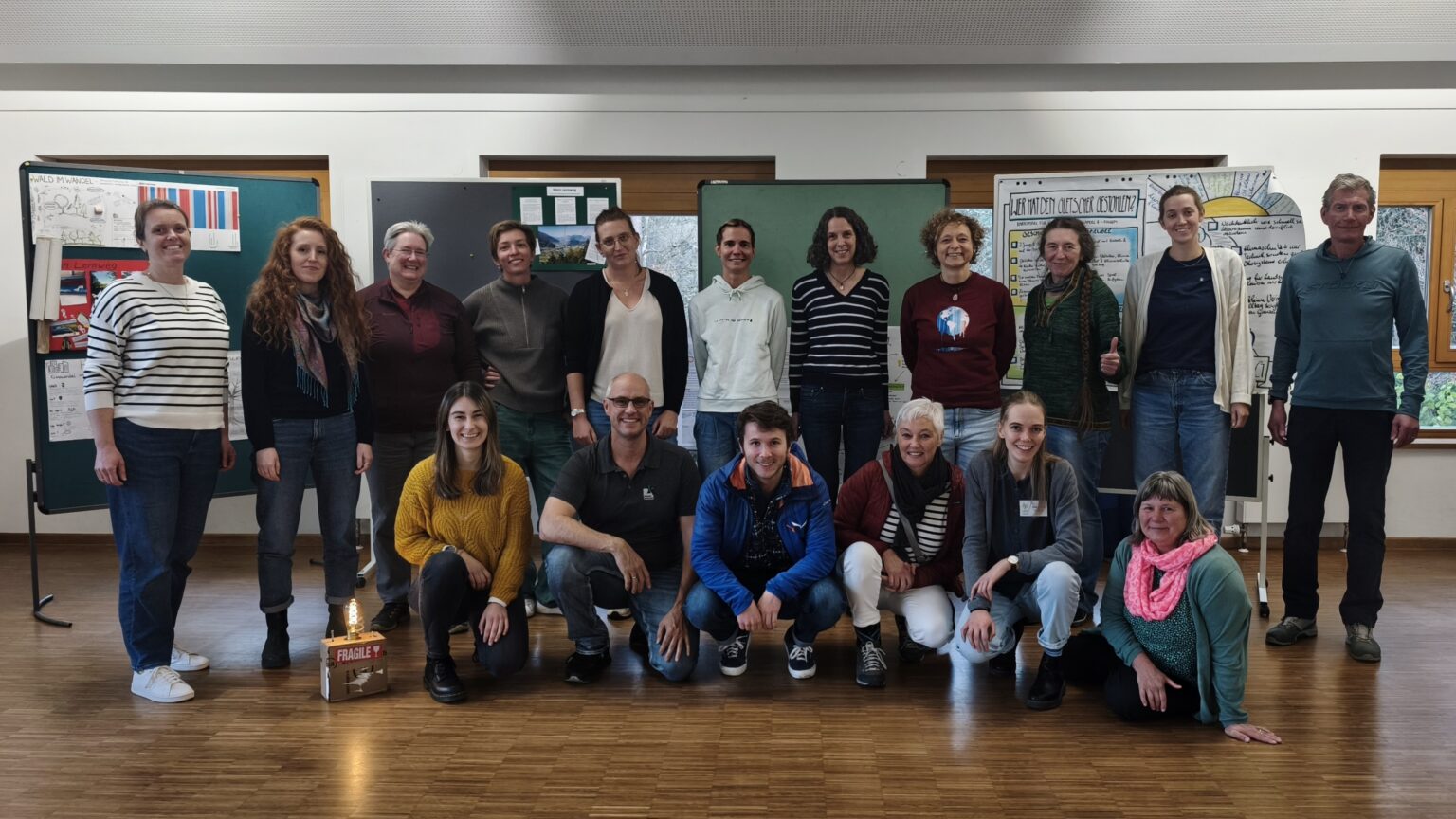 Eine Gruppe von sechzehn KlimaPädagog*innen posiert für ein Gruppenfoto in einem Klassenzimmer mit Holzfußboden, Tafeln und Klimapostern an den Wänden.