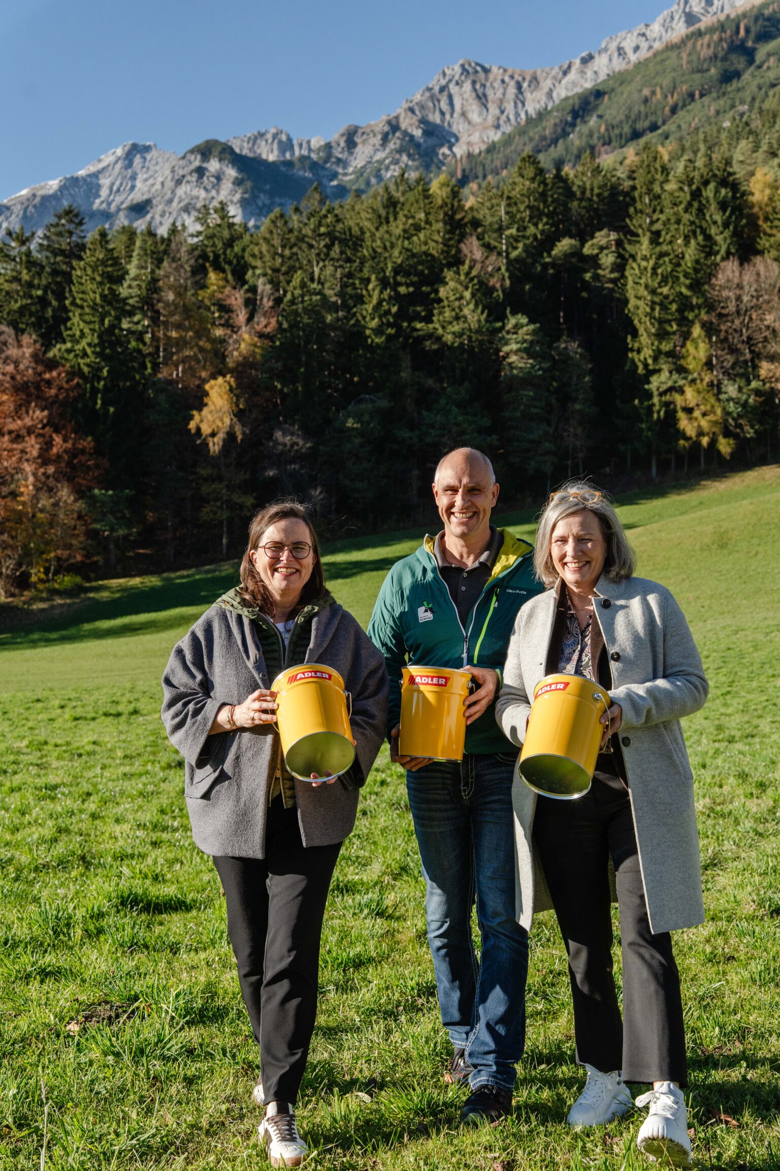 Drei Erwachsene stehen mit ihrem Partner auf einer grasbewachsenen Wiese und halten jeweils große gelbe Becher in der Hand. Ein Wald und Berge bilden die Kulisse unter einem klaren Himmel.