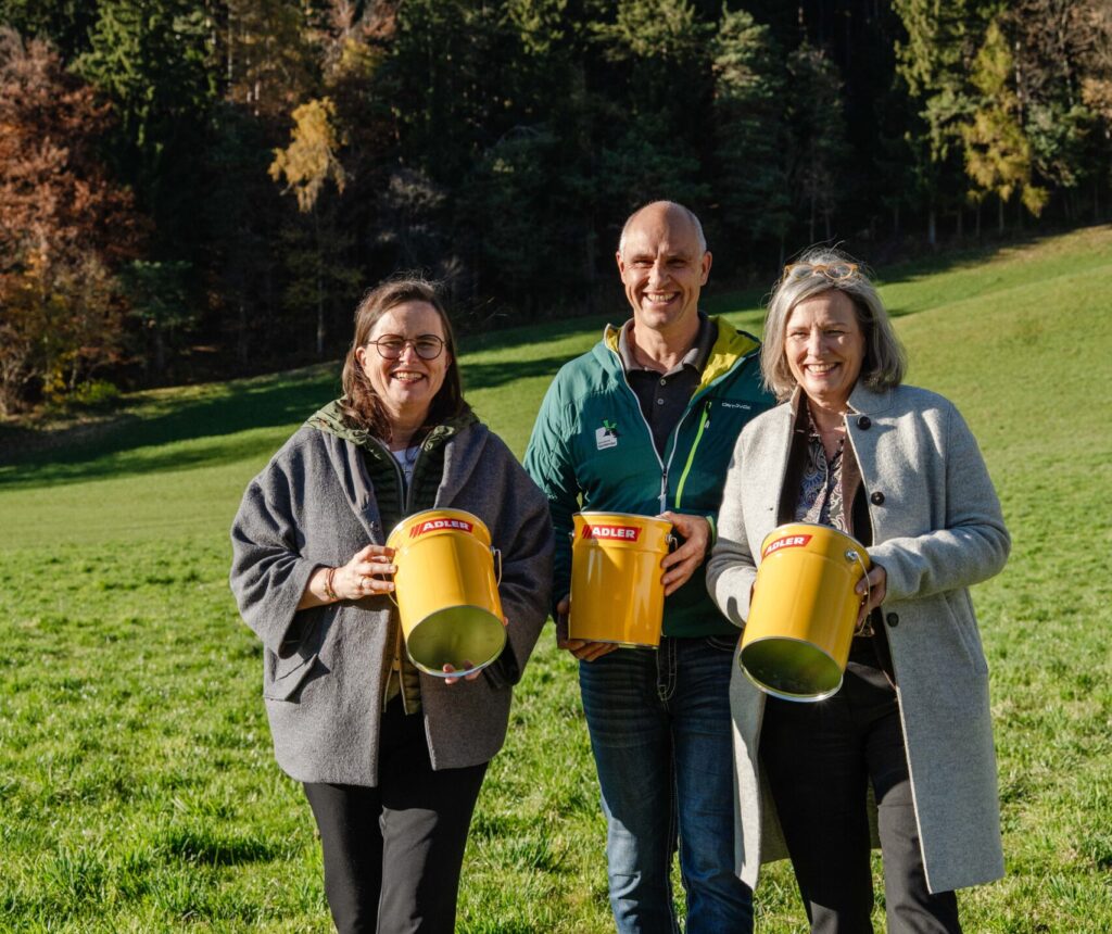 Drei Erwachsene stehen mit ihrem Partner auf einer grasbewachsenen Wiese und halten jeweils große gelbe Becher in der Hand. Ein Wald und Berge bilden die Kulisse unter einem klaren Himmel.