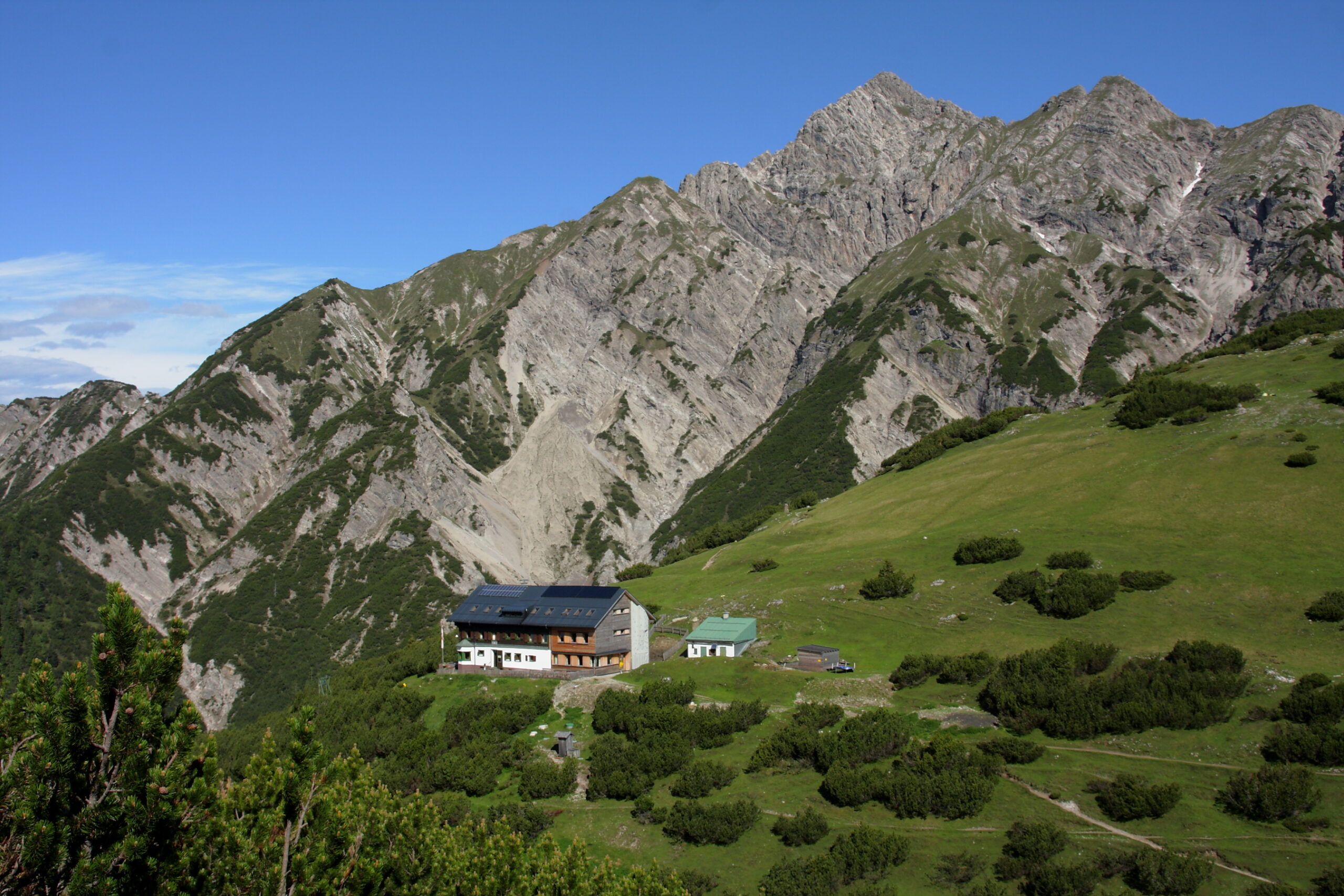Eine Berghütte und ein kleines Gebäude liegen an einem grasbewachsenen Hang und bieten den perfekten Rückzugsort für jeden Junior-Ranger, mit felsigen, steilen Bergen im Hintergrund unter einem klaren blauen Himmel.