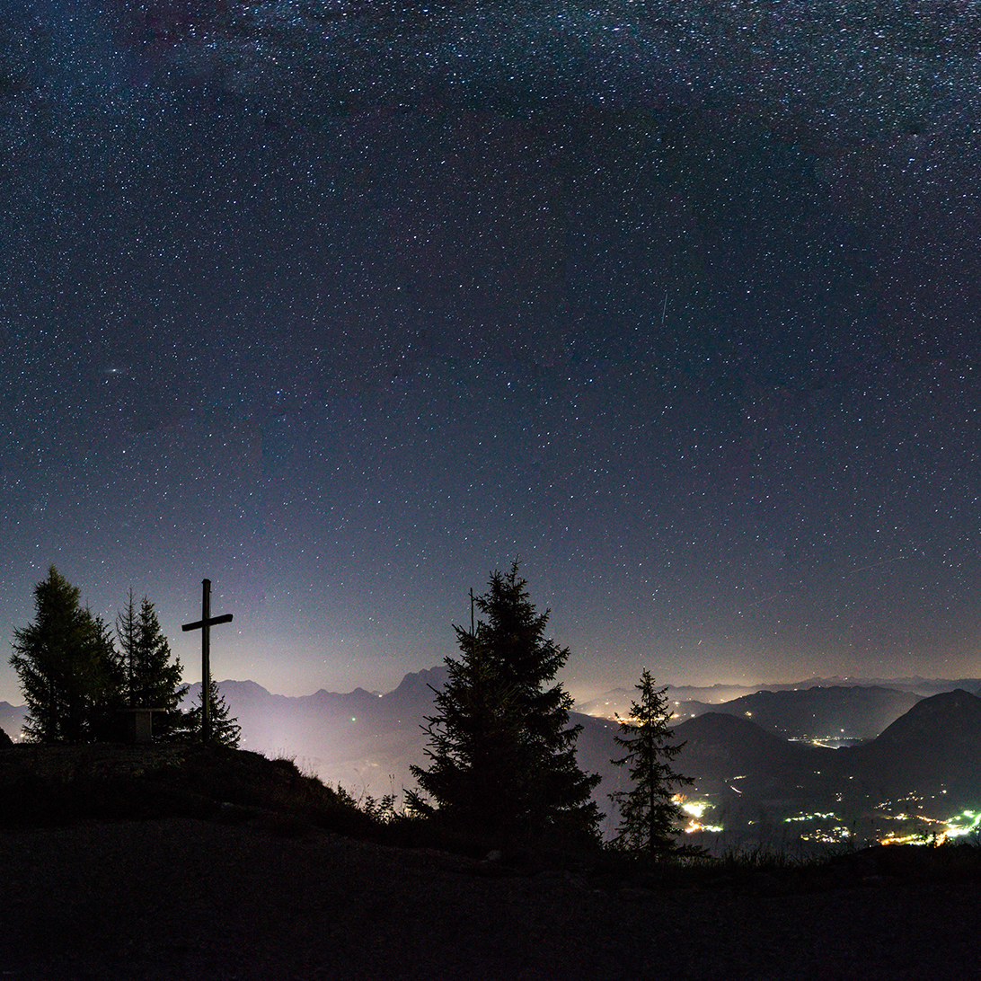 Eine nächtliche Berglandschaft mit einem Kreuz und Bäumen im Vordergrund, die Lichter der Stadt leuchten im Tal darunter und der Sternenhimmel darüber - perfekt für alle angehenden Junior-Ranger, die ein Abenteuer unter den Sternen suchen.