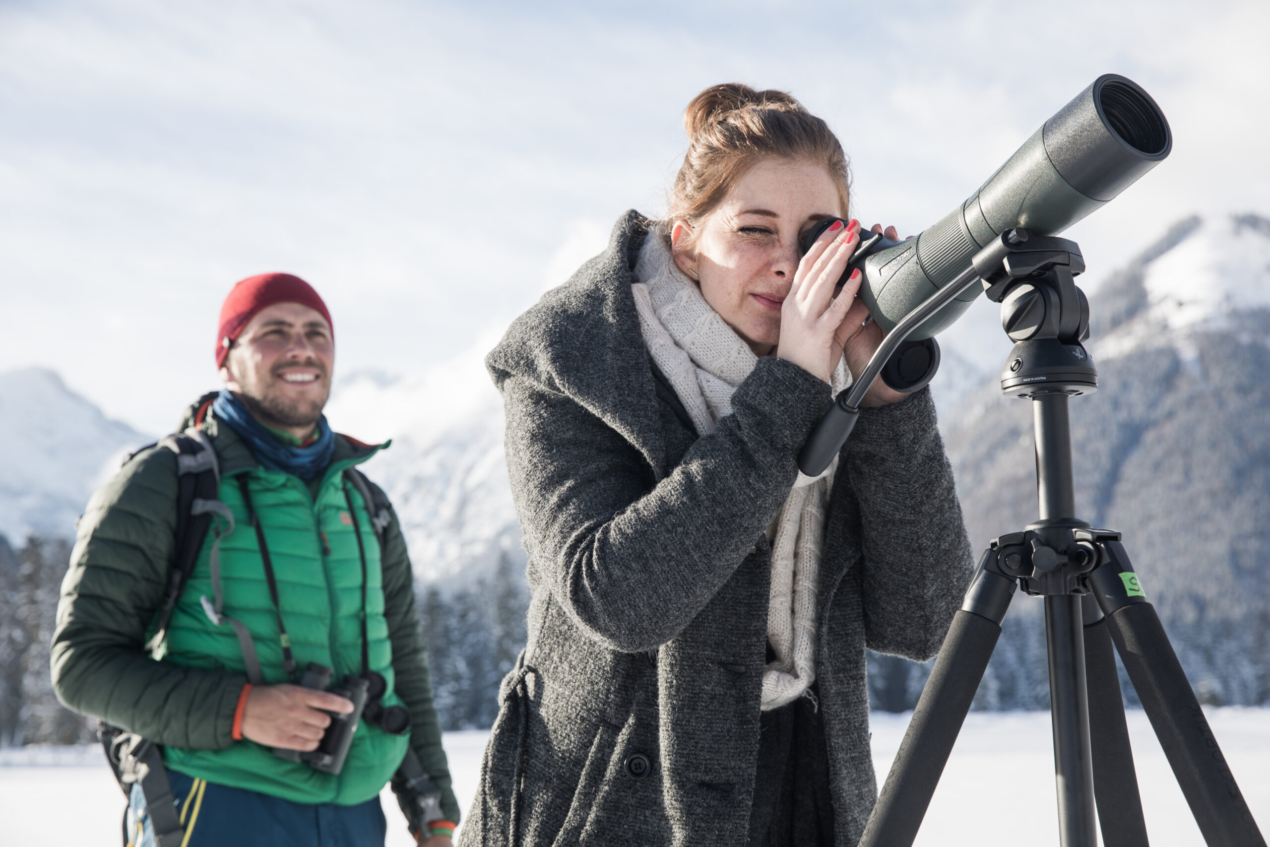Eine Frau schaut während einer Wanderung durch ein Spektiv auf einem Stativ in einer verschneiten Berglandschaft, während ein Mann mit einem Fernglas hinter ihr steht.