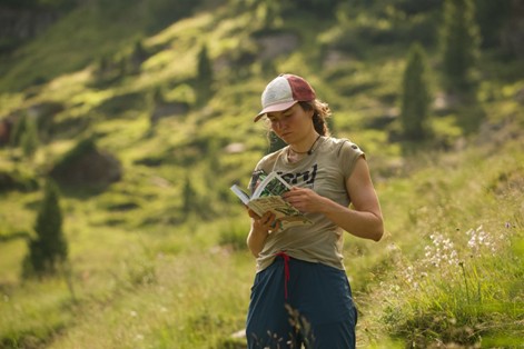 Eine Person mit Mütze und Freizeitkleidung steht im Freien auf einer Wiese und liest ein Buch über aktuelle Forschung, mit Bäumen und Hügeln im Hintergrund.