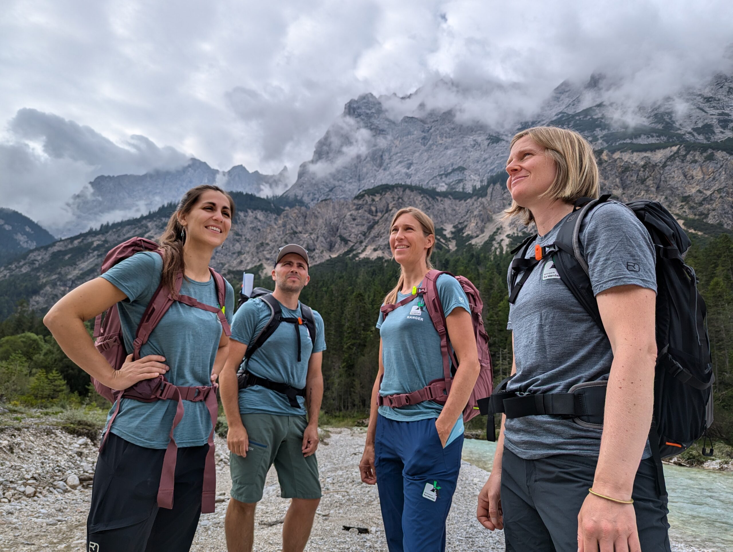 Vier Personen mit Rucksäcken stehen im Freien auf einem felsigen Pfad, umgeben von Kiefern und Bergen unter einem bewölkten Himmel, während ein Naturpark-Ranger den Weg weist.