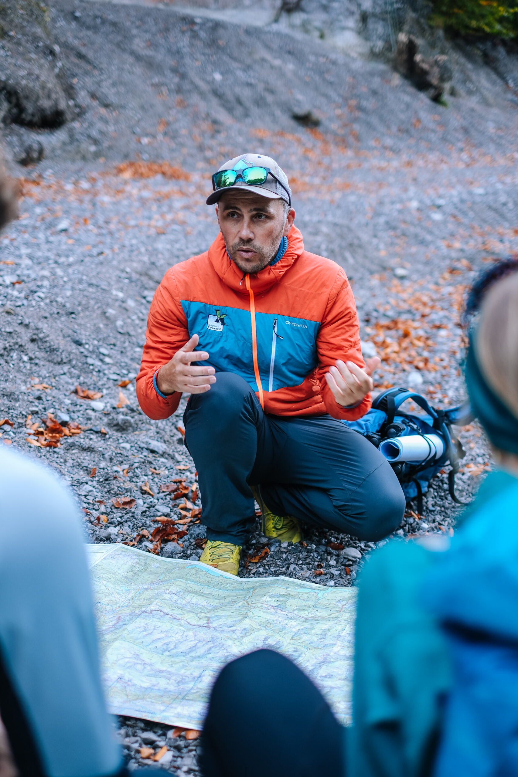 Ein Naturpark-Ranger in Outdoor-Ausrüstung kniet auf felsigem Boden, gestikuliert und spricht mit zwei Personen, während er auf eine vor ihnen ausgebreitete Karte schaut.