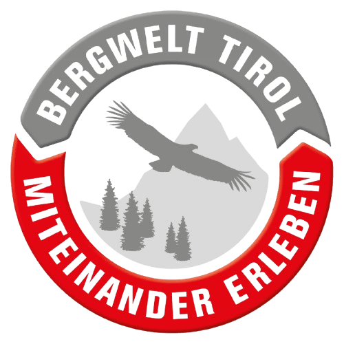 Kreisförmiges Logo mit der Silhouette eines Adlers, der über Berge und Bäume fliegt und an Abenteuer und Natur erinnert. Der Text lautet "Bergwelt Tirol" oben und "Miteinander Erleben" unten, perfekt für Mountainbiken-Fans.