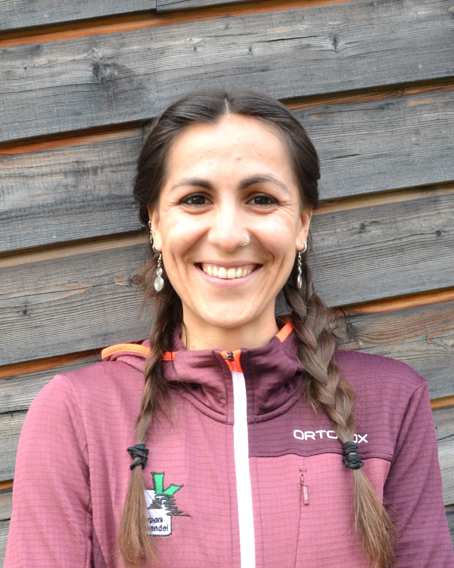 Eine Frau mit geflochtenem Haar und Ohrringen lächelt in die Kamera. Sie trägt eine kastanienbraune Jacke und steht vor einer Holzwand - ihr Stil spiegelt Selbstbewusstsein und Wärme wider und verkörpert den Geist von Klima.