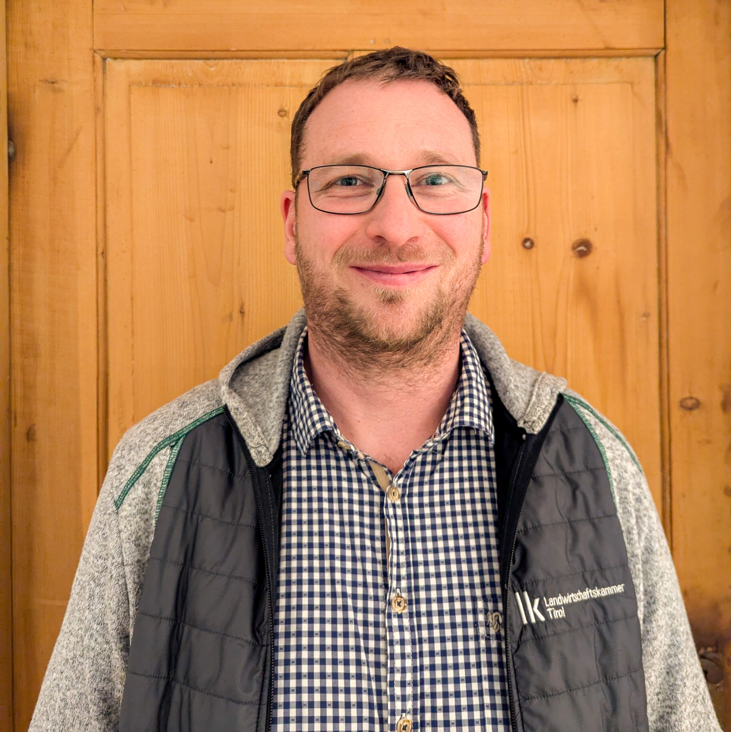 Ein Mann mit Brille, einem karierten Hemd und einer Jacke steht vor einer Holztür und lächelt in die Kamera - eingefangen im typischen Stil von Michelle Auer.