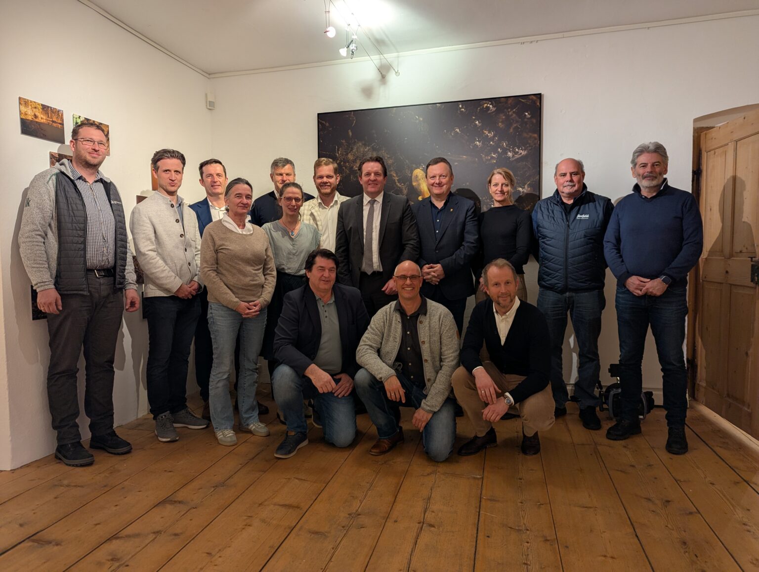 Eine Gruppe von fünfzehn Personen steht in einem Raum mit Holzfußboden und Kunstwerken an den Wänden zusammen. Die meisten stehen, vier knien an der Stirnseite.