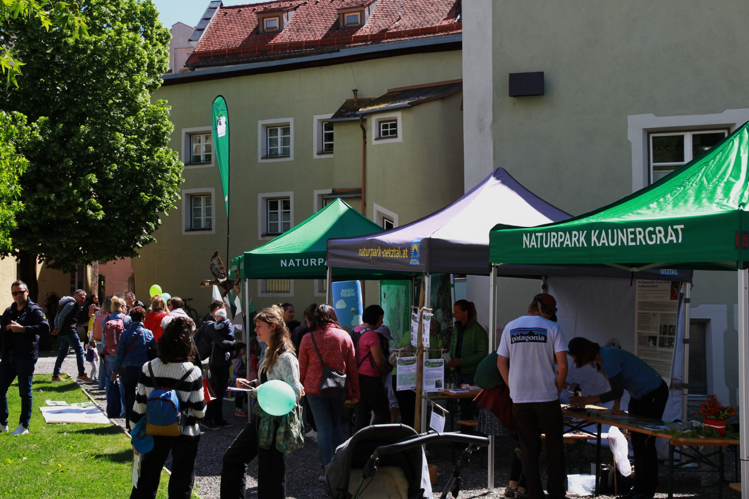 An einem sonnigen Tag versammeln sich die Menschen um Informationsstände mit grünen Zelten mit der Aufschrift "Naturpark Kaunergrat" bei einer Freiluftveranstaltung in der Nähe eines Gebäudes und von Bäumen. Der Naturpark Karwendel zu gast in Hall bringt der Bevölkerung die Natur näher.