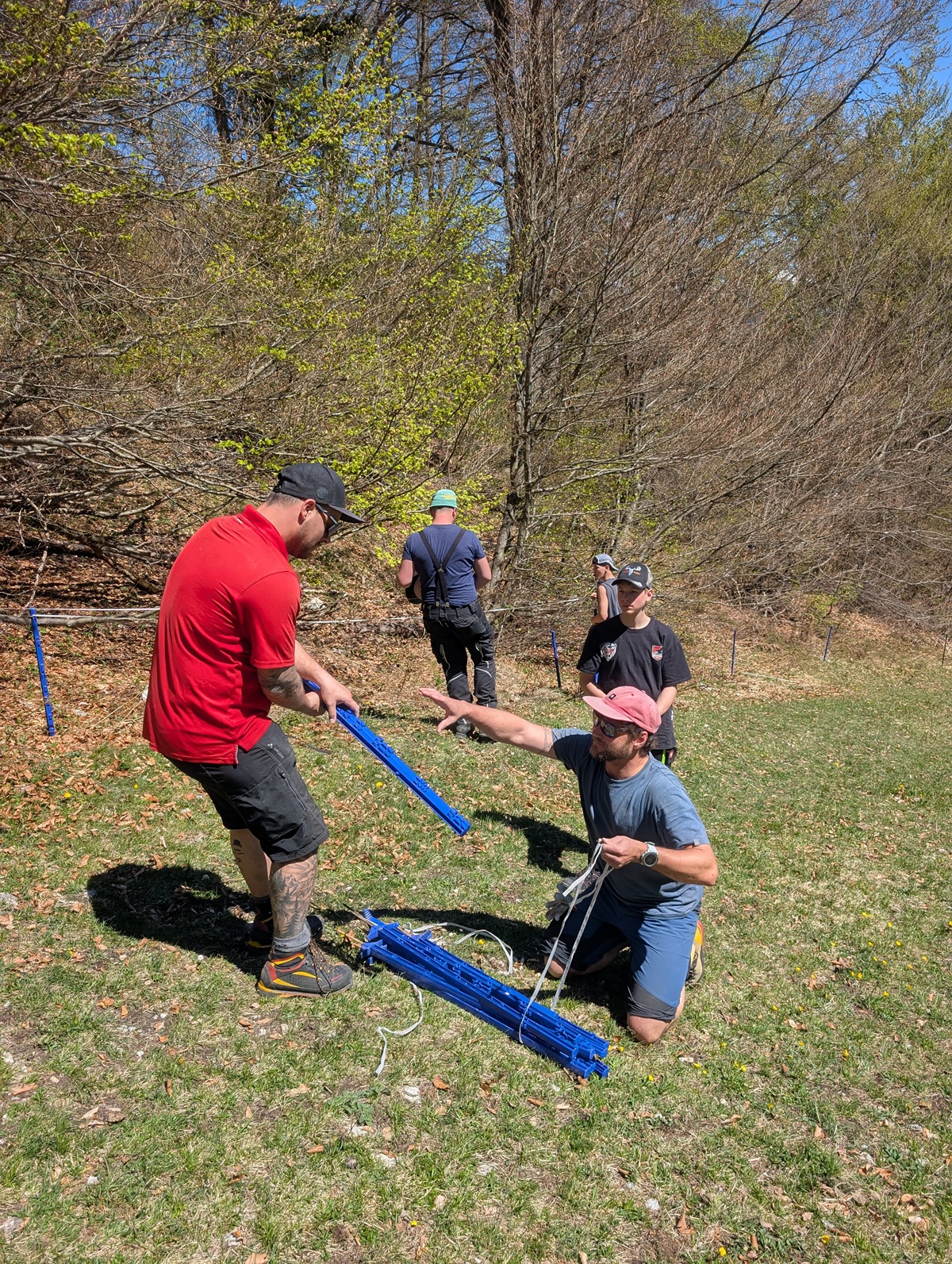 Vier Personen montieren im Freien bei sonnigem Wetter auf einer Wiese mit Bäumen im Hintergrund blaue Plastikstangen und Seile für die Almpflege auf der Arzler Alm.