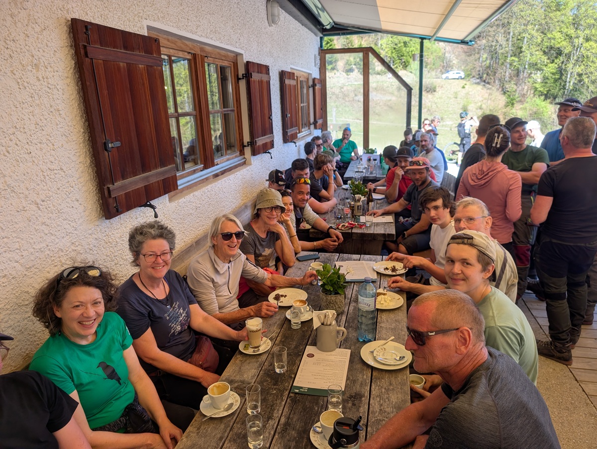 Eine Gruppe von Menschen sitzt an einem langen Holztisch im Freien und genießt Essen und Getränke in der Nähe eines Gebäudes mit hölzernen Fensterläden an einem sonnigen Tag, umgeben von der landschaftlichen Schönheit der Almpflege Arzler Alm.