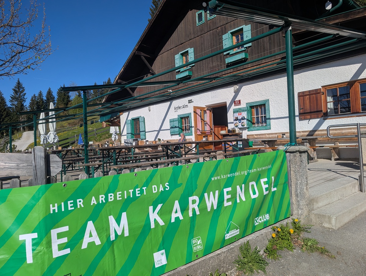 Ein großes grünes Banner mit der Aufschrift "Team Karwendel" hängt vor einem rustikalen Holzgebäude der Almpflege Arzler Alm, mit Sitzgelegenheiten im Freien und einem klaren blauen Himmel darüber.